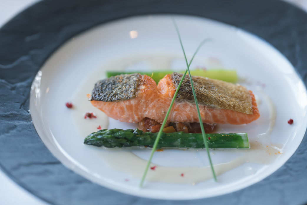 大きな窓から自然光が差し込む明るいレストラン会場。テーブルには美食とワインが並び、ゲストが景色と食事を楽しむ様子